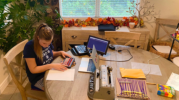 A white high school student sits at her kitchen table preparing to work a problem on her Cranmer abacus. Her braille notetaker, laptop, Perkins braille writer and other materials are visible on the table.