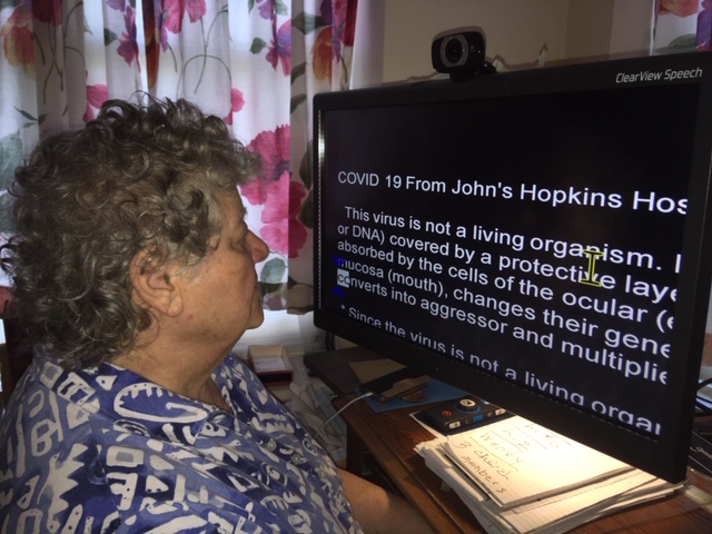An older woman reads information about the COVID-19 virus using a desktop video magnifier (CCTV). The font on the screen is white and the background black.