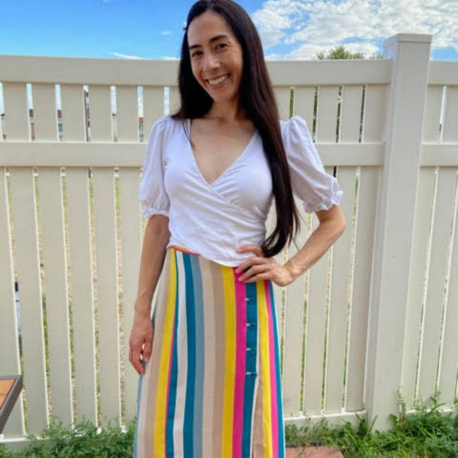 Elizabeth Bolander stands smiling in front of a white fence with her hand on her hip. She is wearing a white V-neck shirt and a long multicolor-striped skirt. 