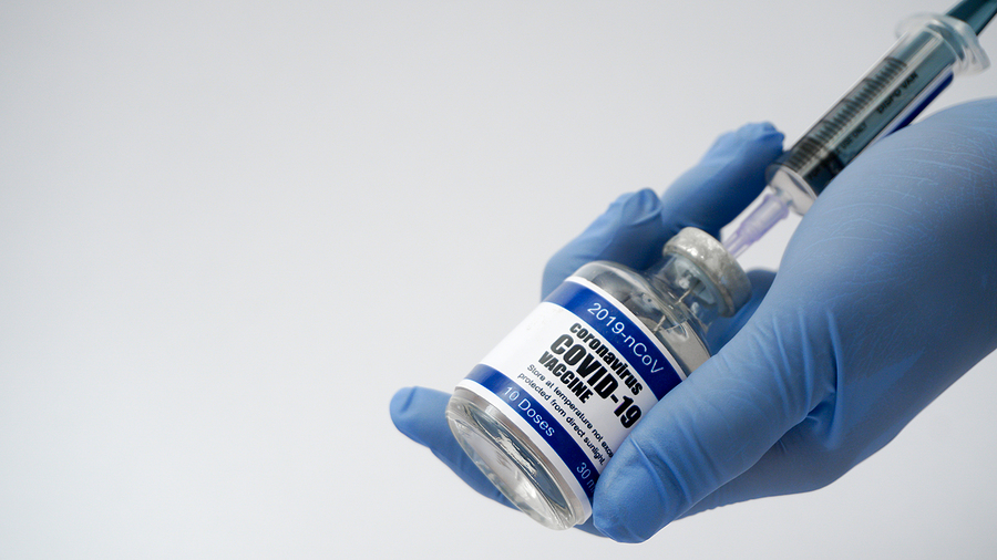 latex-gloved hand holds a vial of vaccine as another gloved hand inserts a syringe into the vial.