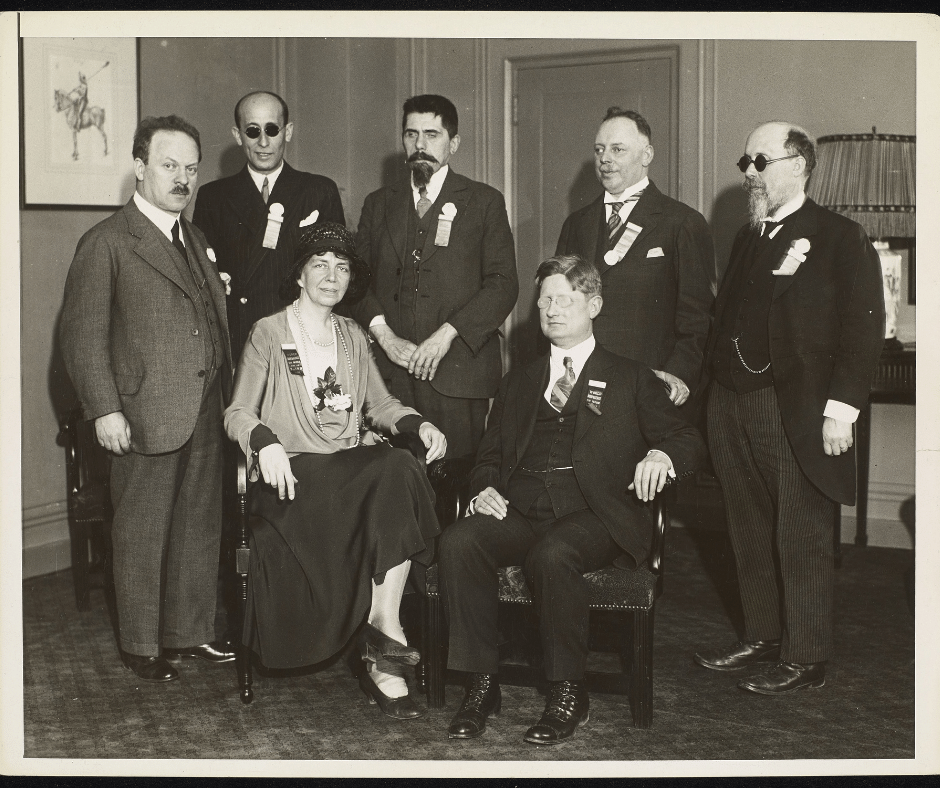 Five men, four of whom appear blind, stand behind Eleanor Roosevelt and Robert B. Irwin who are seated infront at the World Conference on Work for the Blind. They are pictured in one corner of a room with a framed image behind them on one wall, and a door and table and a lamp behind them on the other. Roosevelt is wearing a bucket type hat and very loose clothing.