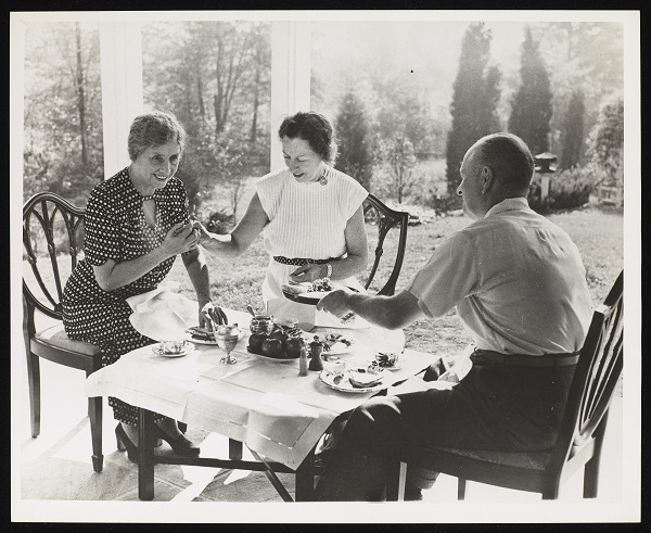 Taken on a covered porch looking out onto Helen Keller's garden in Westport, Connecticut. Helen Keller, Polly Thomson and Herbert Haas are seated left to right around a small square table set with food. Keller and Thomson are communicating using manual sign language. Keller wears a short-sleeve dress with a design of small horizontal dots and flowers. Thomson's light color dress has thin vertical piping, cap sleeves and is belted at the waist. The image is a still from the film "Helen Keller in Her Story."