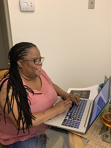 A woman with brown skin, long braids, and glasses with thick lenses and a big smile sits at a laptop computer