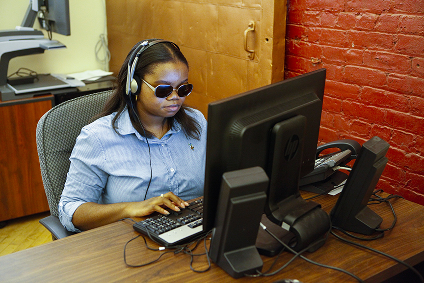 A woman with brown skin wearing dark glasses and headphones sits at a computer