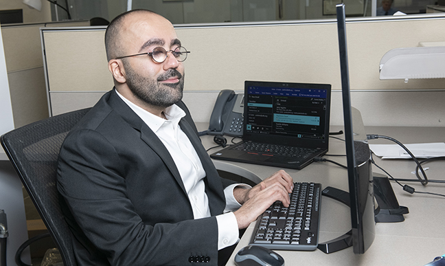 A man with light brown skin, wearing a suit jacket and glasses with thick lenses, sits at a desktop computer with his hands on a keyboard