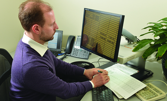 A professionally dressed man with light skin and brown hair writes under the camera of a CCTV as he views the magnified text on the screen