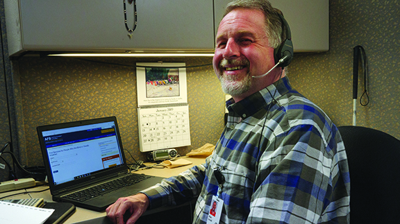 A smiling man with light skin and graying hair and beard wears a headset and sits at a laptop computer in an office cubical with a long white cane propped against the wall