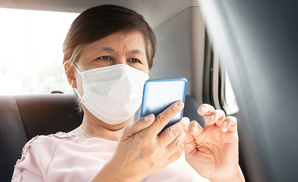 A person with light skin wearing a face mask and seated in the back seat of a car is swiping the screen of a cell phone