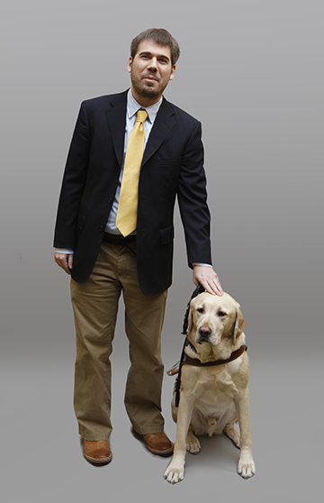 A smiling man in suit and tie is petting his guide dog on the head