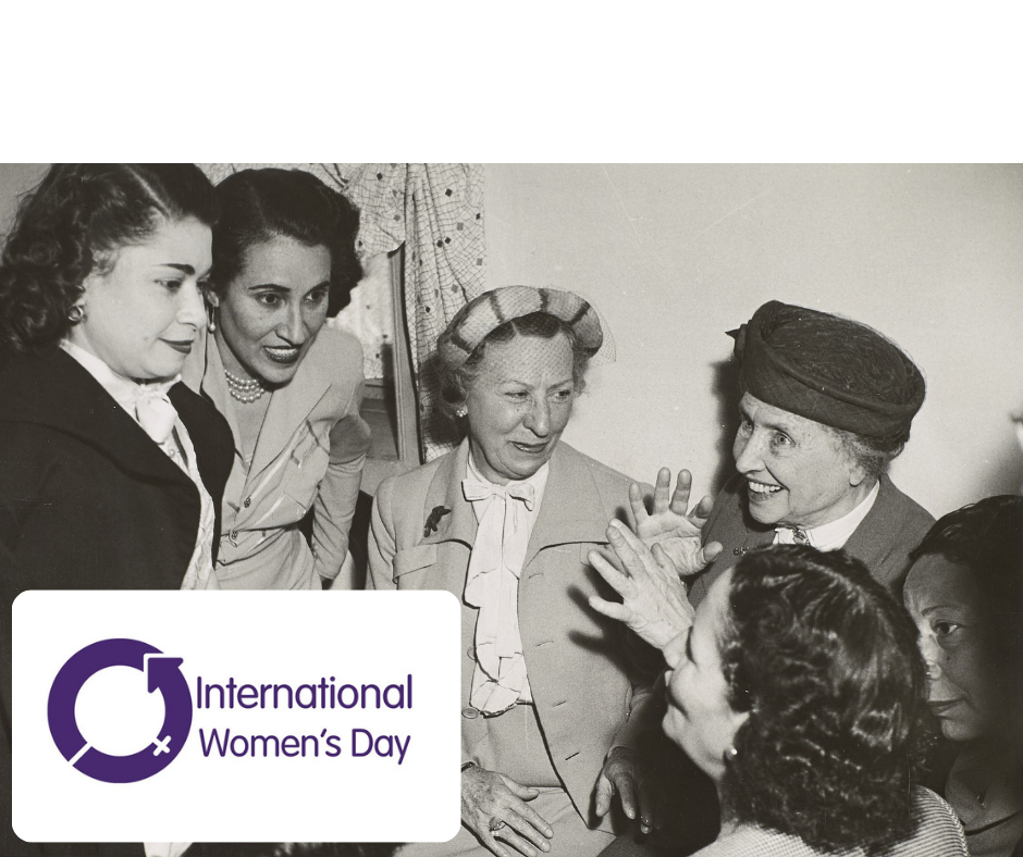 Logo: International Womens Day. Image Description: Taken indoors during Helen Keller's visit to the "School for blind girls" Aboukeer, Alexandria, Egypt. Close-up image of Polly Thomson and Keller seated side-by-side and left to right. They are circled by "distinguished" female members of the "Welfare of the Blind." Keller is smiling broadly and her hands are raised as she speaks. Two women can be seen seated to her left (bottom right of the image), while three women are seen standing on Thomson's right.
