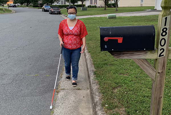 A woman is walking down a residential street. She is wearing a mask and holding a white cane. She is also wearing glasses and a red shirt. She is in the process of walking by a residential mailbox which is located on the left side of the photo.