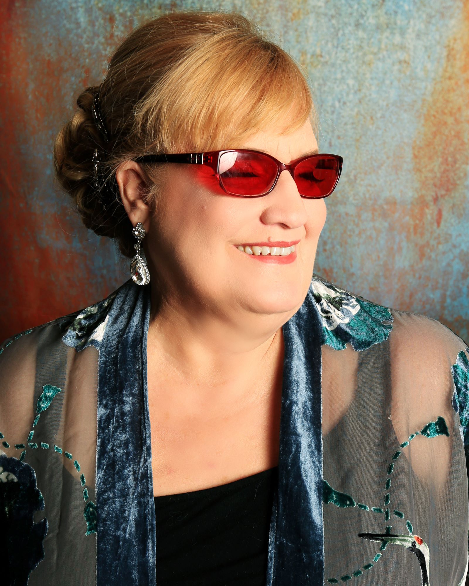 Alice smiling, wearing shades and fish-shaped earings. Dark blonde hair pulled back and wearing a blouse.