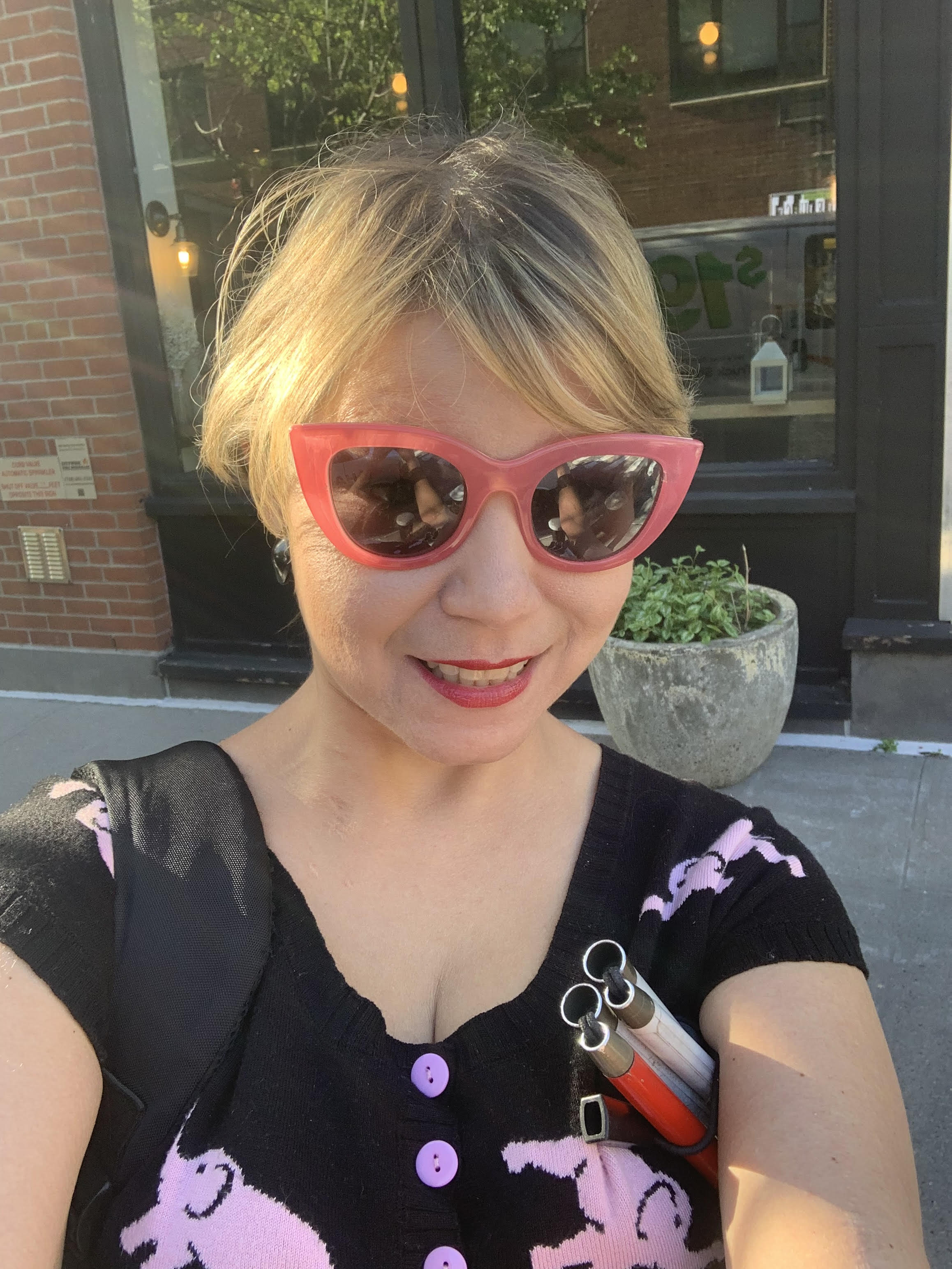 Bojana, a white woman with blonde bangs combed forward, smiling broadly. The photo captures a sense of joy and springtime, as the sun is shining onto my face. I am wearing sunglasses with pink frames, bright pink lipstick and I am wearing a black cardigan with floating pink elephants printed on it.