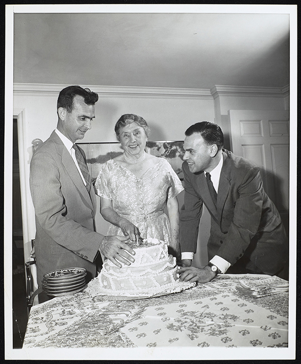 Helen Keller cutting a 3-tiered cake on her 75th birthday at her home in Westport, CT. The cake is being presented by M. Robert Barnett (AFB) who is on her right and Eric T. Boulter (AFOB) who is on her left. A single candle is on the cake and Barnett and Boulter both touch the cake.