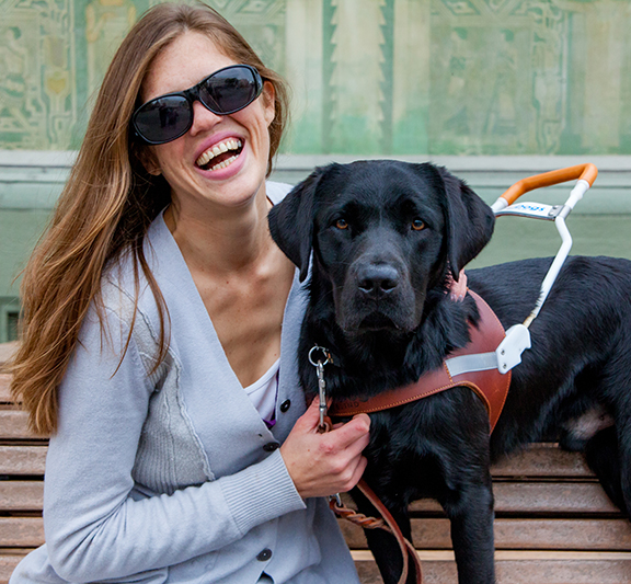 A smiling woman sits on a bench next to a black Lab guide dog.