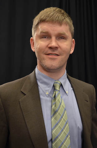 A white middle aged man, wearing a blue shirt and a grey suit. He is smiling. 