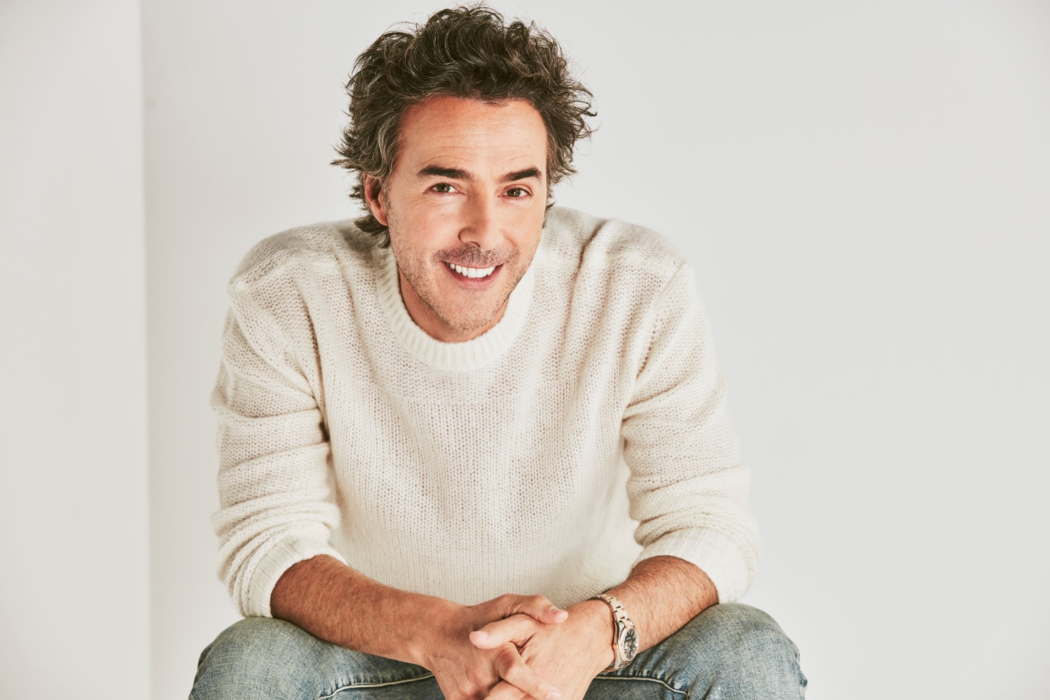 Shawn Levy, a man with shaggy brown hair and a vibrant smile, leans forward as he sits with his arms resting on his legs.