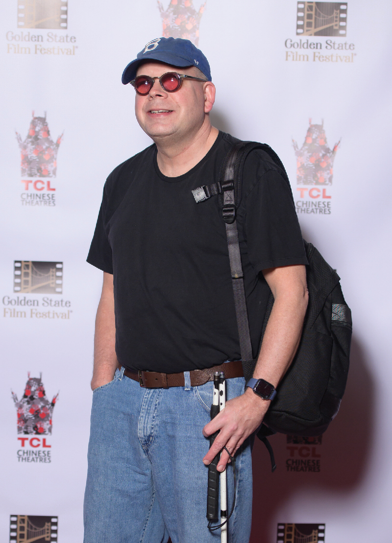 A smiling man with a white cane stands in front of a logo backdrop.
