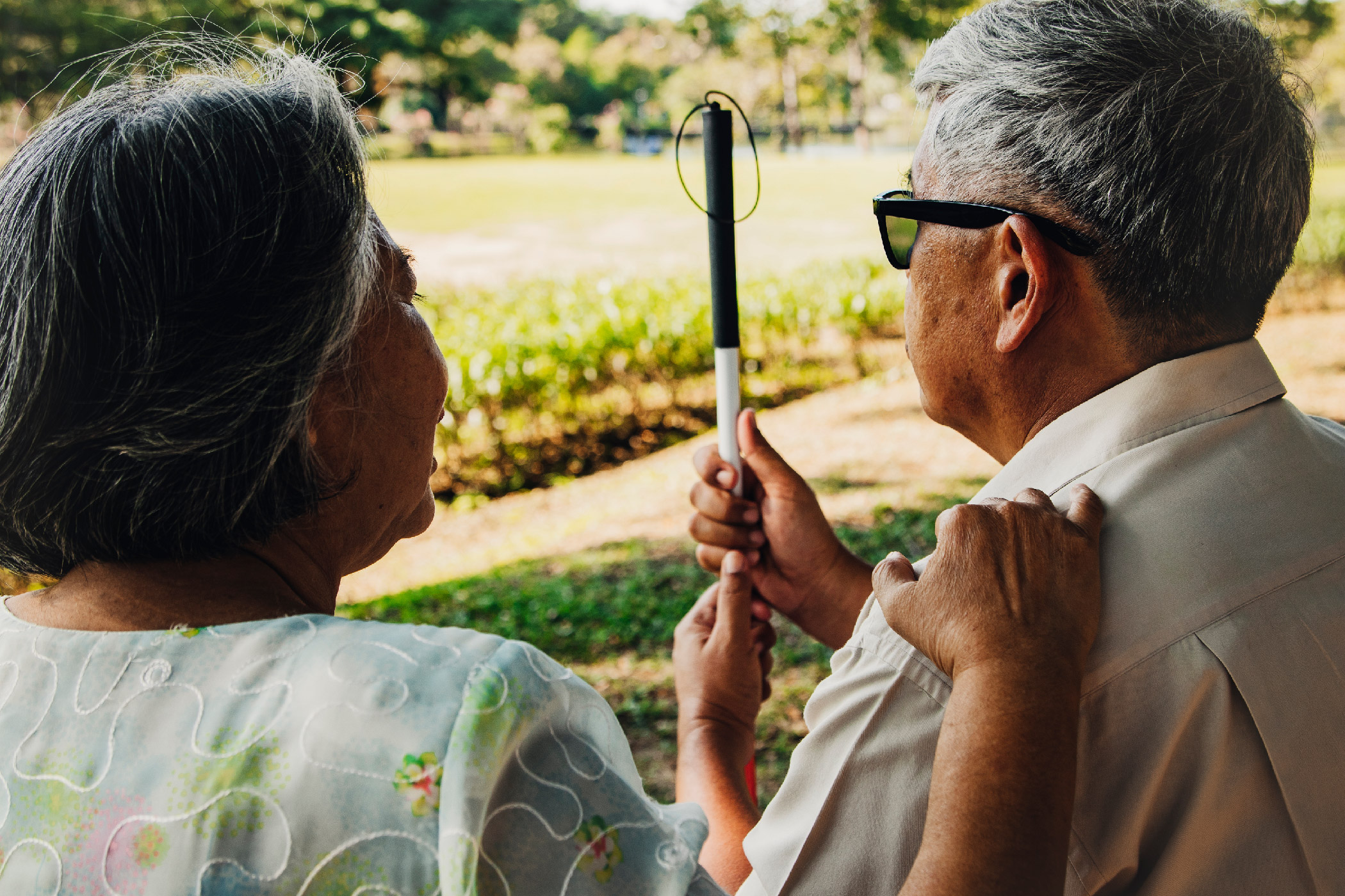 An older woman and man with a white cane sit in a park.