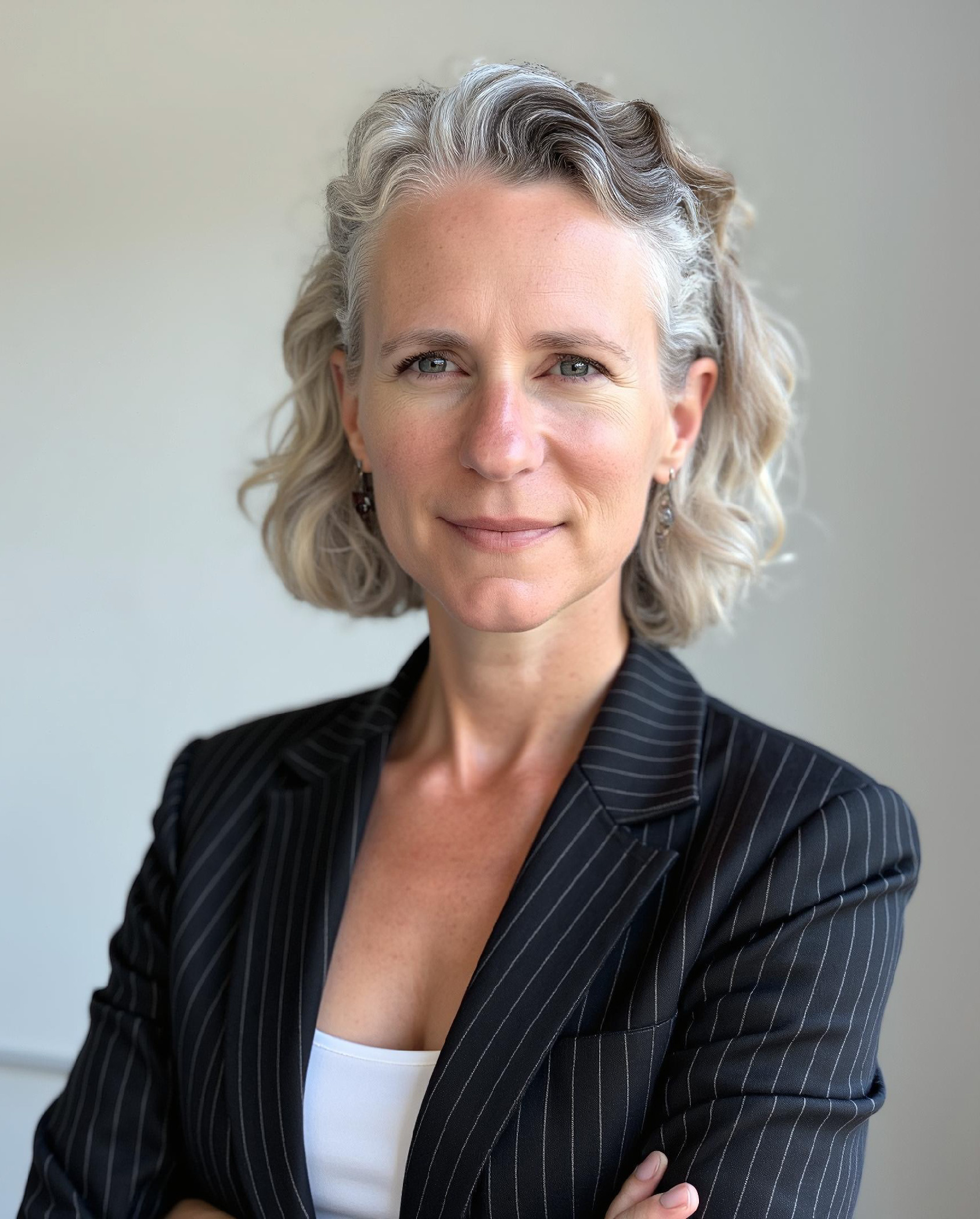 A woman with curly chin-length silvery blonde hair wears a sophisticated black blazer with thin white stripes.