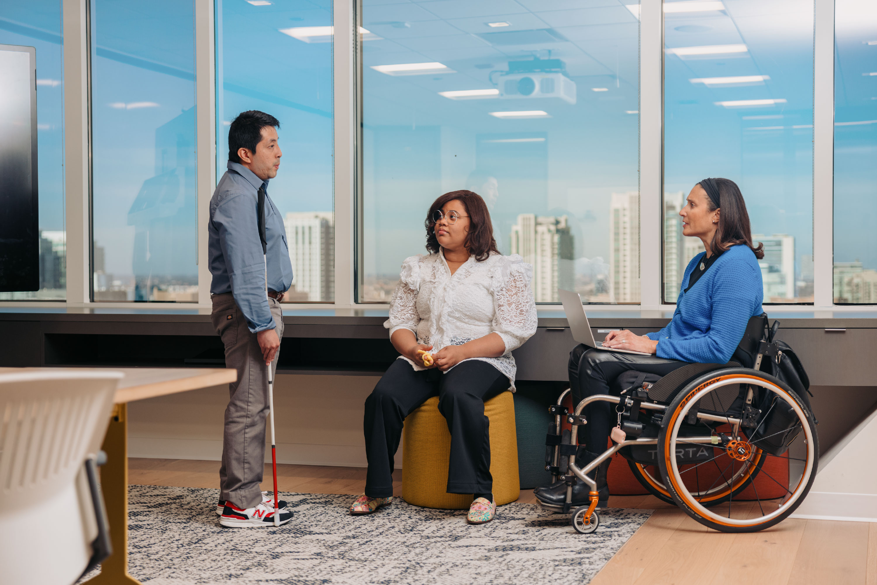 Three colleagues converse in a meeting room. One stands with his white cane in one arm. Beside him, a young woman sits holding a fidget toy next to a woman who is using a wheelchair.