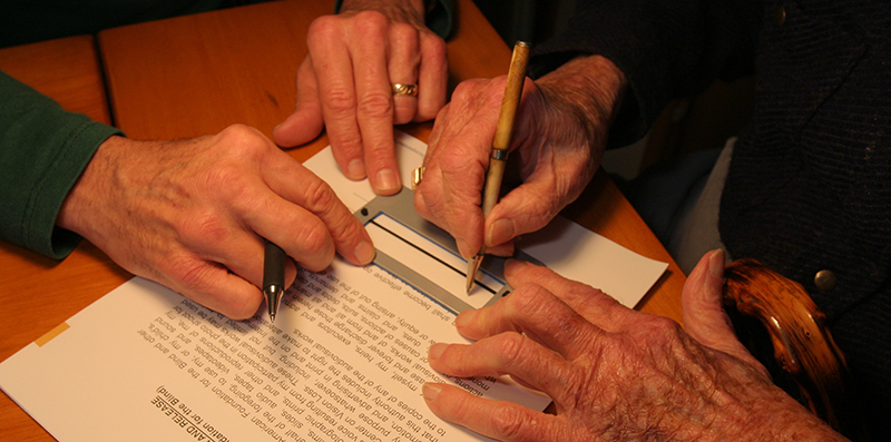 Closeup of an older person using a small plastic card with a rectangular cutout to guide their signature on paper. Another person's hands are on the guide, to ensure that it's in the right place.