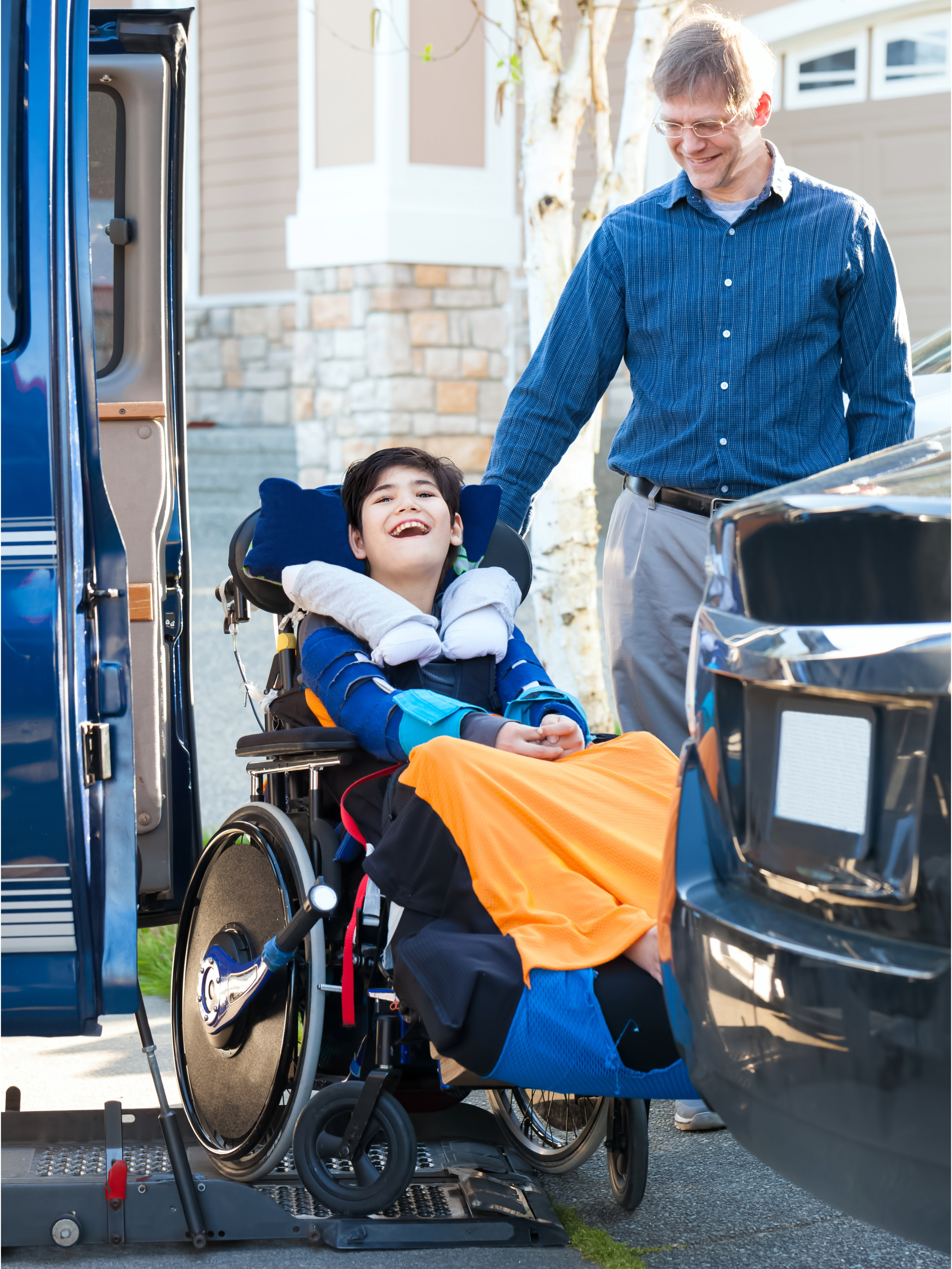 A smiling man stands beside a smiling child in a wheelchair.