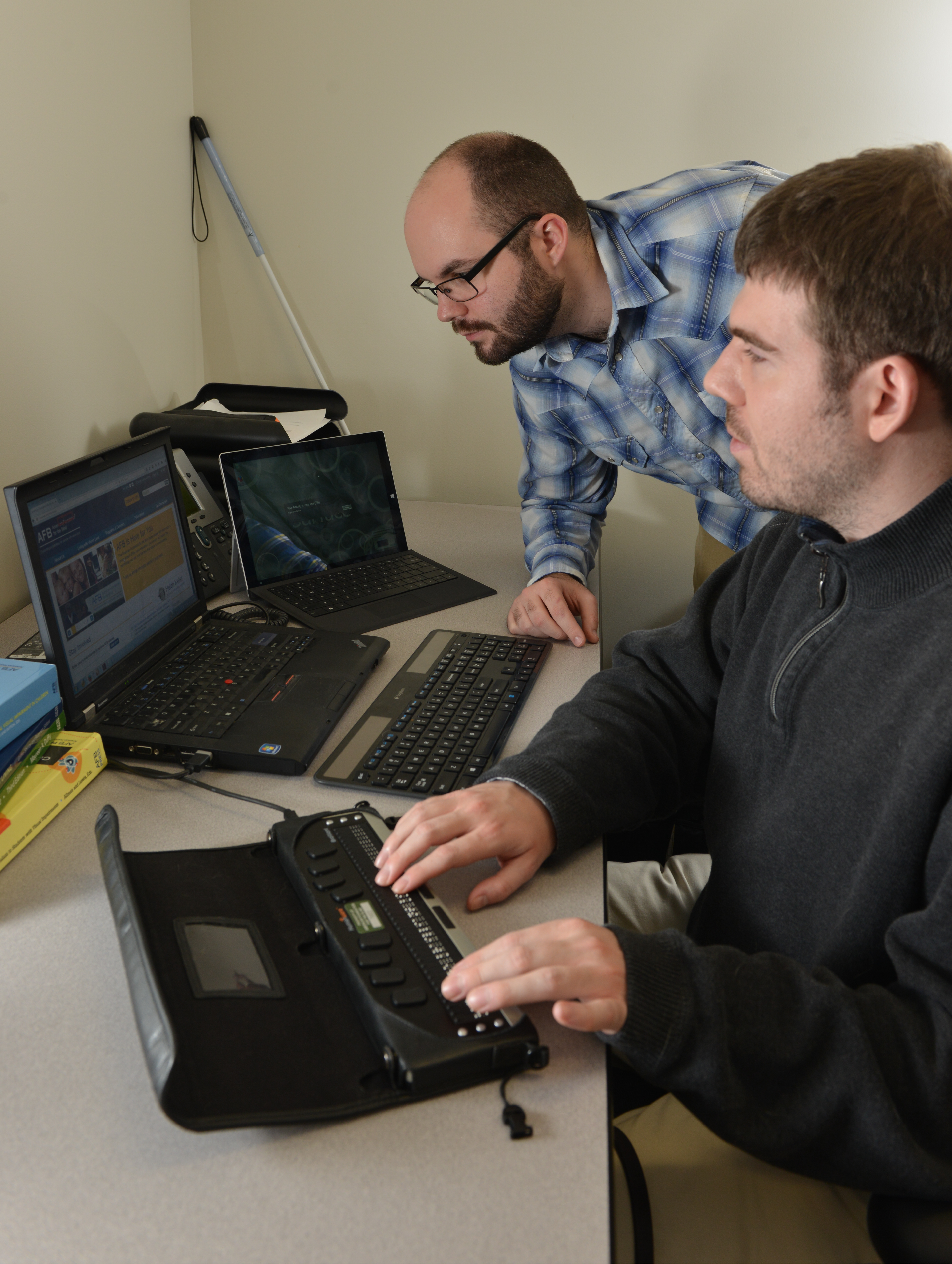 A man types on a braille keyboard while another man beside him looks at a computer screen.