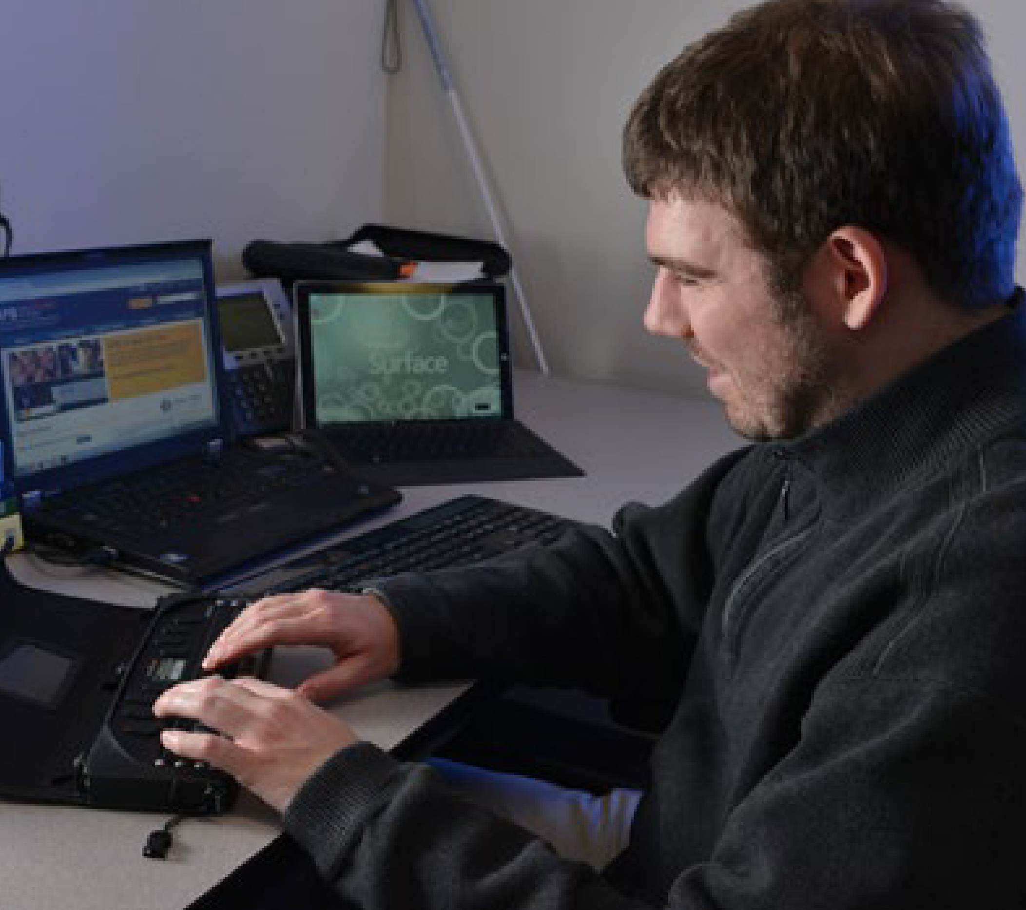 A smiling man types on a braille display connected to a laptop.
