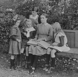 Tilly Aston in her 20s, sitting outside on a bench reading braille and surrounded by small children