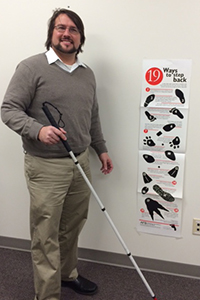 Joe Strechay with white cane in hand, posing by the 19 Ways to Step Back poster