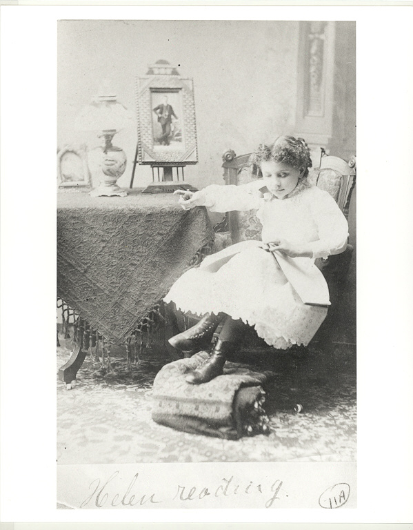 Helen Keller reading a braille book with her left hand and signing the shape of a letter with her right. At the Perkins School for the Blind, 1888