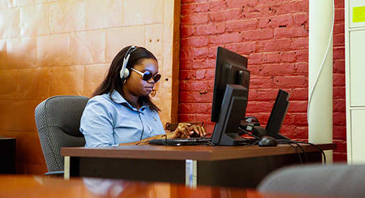 woman sitting at work desk, wearing sunglasses and using computer with headphones