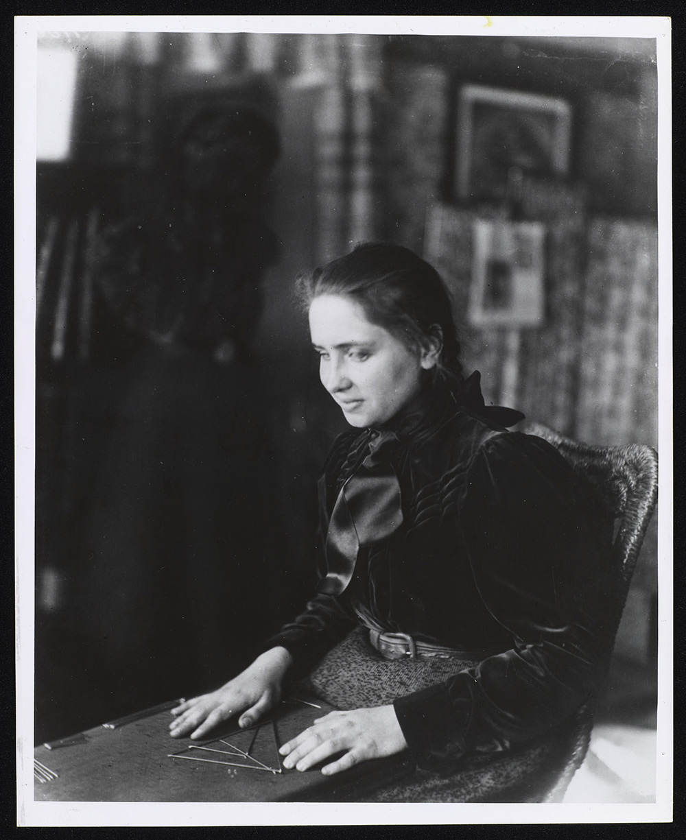 Helen Keller is seated alone studying geometry at Radcliffe College. Keller has a board on her lap and her hands are touching rods that are arranged in a triangular geometric pattern. She is wearing a dress that appears to have a dark satin-like upper body, with a wool-type fabric bottom part.The outfit has sleeves that are tight from wrist to elbow and puffy from elbow to shoulder. The flouncy collar reaches high up her neck. Her hair is lightly pulled back into a single braid with a bow just barely visible. 