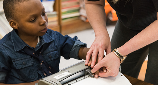 A boy sitting at a desk using a braille writer with help from his teacher