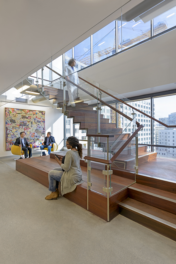 A man in a white suit walks down the stairs in the Lighthouse entryway, holding the handrail and looking out the large windows. A woman is checking her phone, sitting on the wide wooden bench extending from the foot of the stairs. Two men converse in chairs positioned in the light-filled seating area behind her.