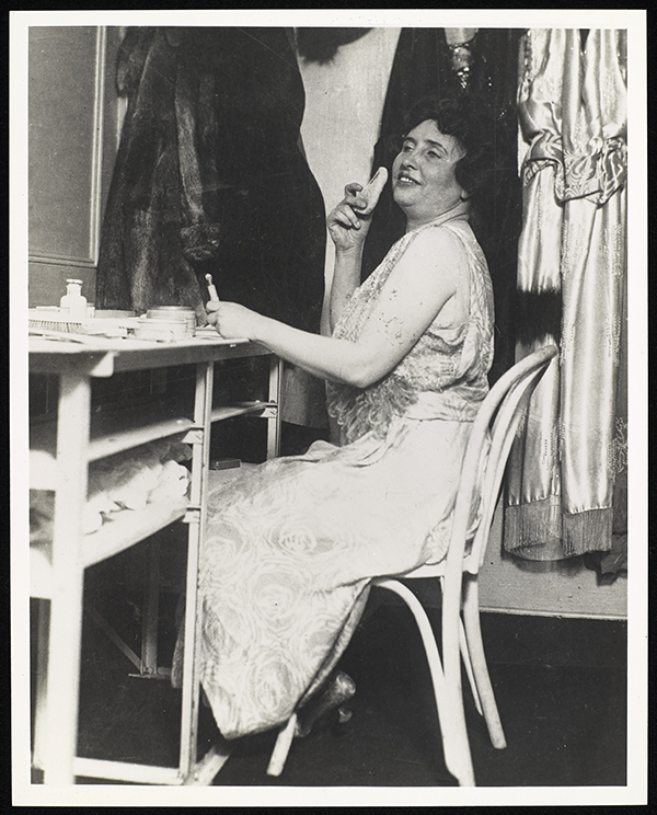 Helen Keller in her vaudeville dressing room. She is seen sitting with her head tilted slightly back as she faces slightly towards the camera. She is applying powder with her right hand, while in her left she holds a cosmetic item. Her left hand is leaning on a table in front of her that is covered with other make-up items. She wears an ankle length, sleeveless shimmering dress that is loose except at the waist. A fur coat and silk or satin items of clothing are visible behind her.