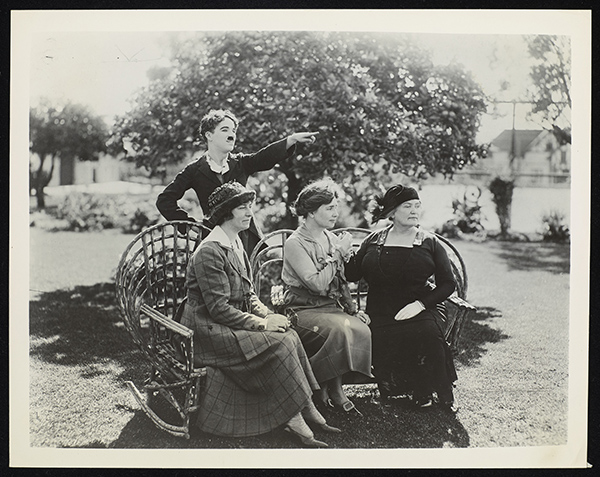 Viewed outdoors, Charlie Chaplin stands behind Polly Thomson, Helen Keller and Anne Sullivan Macy who are all seated on wicker chairs. Their faces are seen in profile as they look towards the right of the image, in the direction that Chaplin is pointing. The women's skirts and dresses are long. Thomson and Macy wear hats. Macy is manually signing into Keller's right hand.