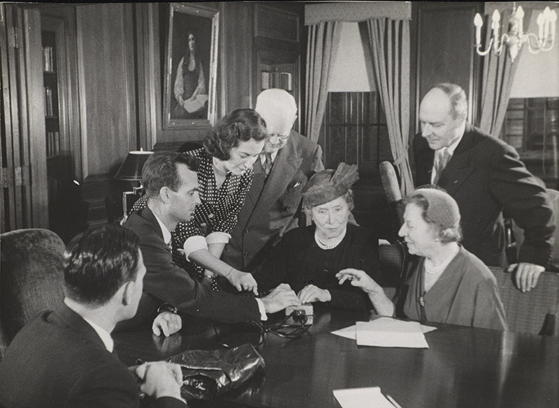 Left to right are: unidentified man, M. Robert Barnett (AFB Executive Director), Marta Sobieski, Peter J. Salmon (Executive Director, Industrial Home for the Blind, Brooklyn), Helen Keller, Polly Thomson, and Gregor Ziemer. The room has wood paneling and a painting of Helen Keller by Albert H. Munsell is visible in the background. Image from documentary Helen Keller in Her Story.