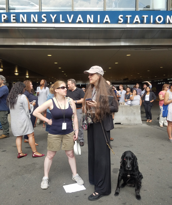 Alina Vayntrub and Crista Earl stand outside Penn Station, with Crista's dog guide Paige. Alina holds a colander and Crista, wearing headphones, is holding her iPhone. A piece of white paper lies on the ground.