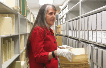 A researcher standing in the aisle of the Helen Keller Archive, examining a braille book. She is wearing white gloves, to protect the pages, and her white cane is tucked under her elbow.