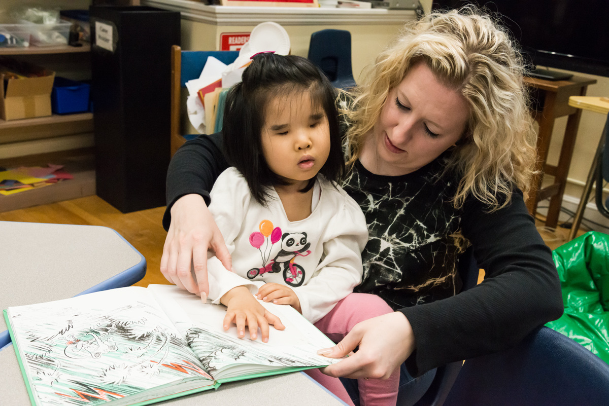A teacher helps a young girl read a braille book.