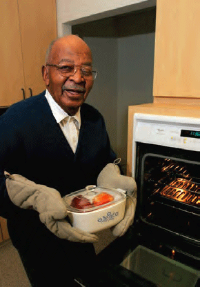 Older man standing by an oven, using hotpads to hold a casserole. He looks pleased.