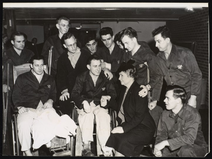 Taken during Helen Keller's tour of U. S. Military Hospitals. Keller is seated in a ward at Vaughan General Hospital, Hines, Illinois. She is with ten veterans. Most of the men are standing behind two veterans in wheelchairs. Keller, who is seen in three-quarter profile (facing the left-hand side of the image), is holding the hand of the veteran in the wheelchair who is to her right and slightly in front of her. Also to her right and ahead of her is the second man in a wheelchair, an amputee. Many of the m