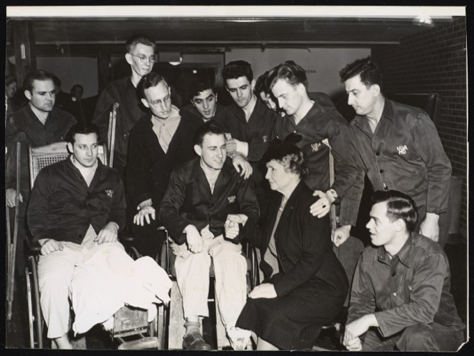 Taken during Helen Keller's tour of U. S. Military Hospitals. Keller is seated in a ward at Vaughan General Hospital, Hines, Illinois. She is with ten veterans. Most of the men are standing behind two veterans in wheelchairs. Keller, who is seen in three-quarter profile (facing the left-hand side of the image), is holding the hand of the veteran in the wheelchair who is to her right and slightly in front of her. Also to her right and ahead of her is the second man in a wheelchair, an amputee. Many of the men wear military overalls.Three men are on crutches. One of the men on crutches has his left hand on Keller's shoulder. Keller is wearing a dark pinstripe jacket and skirt, a light-color top and a dark hat.