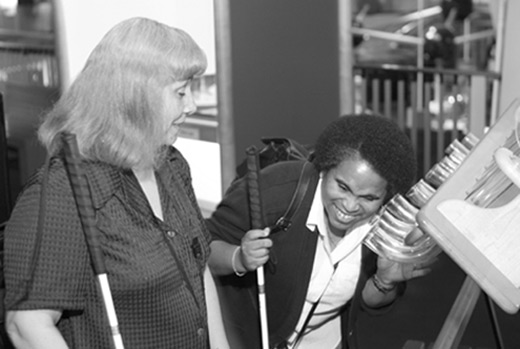 Figure 2: Photograph of two women with long canes, one listening at the open end of a tube that is part of a bank of a number of clear, curved tubes and one watching.