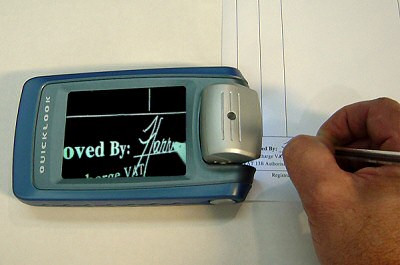 Photo of the QuickLook on a piece of paper. To the right is a hand holding a pen over some text. The screen of the device displays, in reverse polarity, "...oved By:" and the beginning of a signature "Harr..." and the tip of a pen.