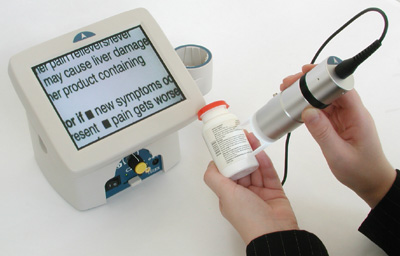 Photo of hands holding a cylindrical device with two white prongs against a medicine bottle. The separate monitor display, in large print, reads "...her pain relievers/fever...may cause liver damage...her product containing...or if new symptoms oc...esent...pain gets worse..."