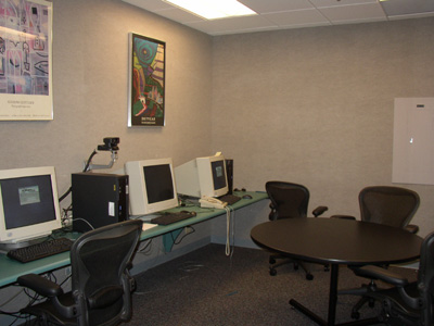 Photo of a room with several computer monitors, keyboards, and other equipment, desk chairs, and a small round table.
