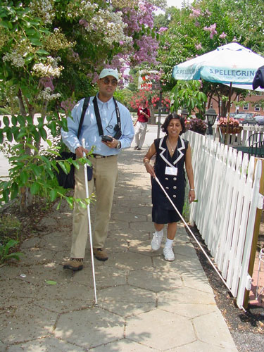 A tall man and a short woman, both with canes, are walking down a sidewalk past an outdoor café. The man holds the Trekker, which is attached to a strap worn around his neck.